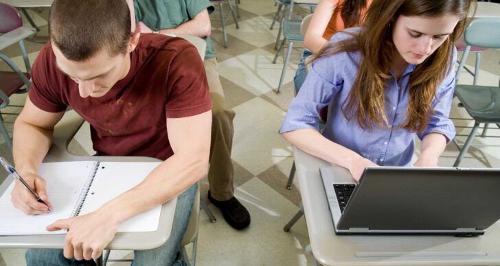 students in class - one using a laptop and one writing in a notebook