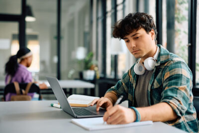 Young student taking notes while e-learning on laptop at school.